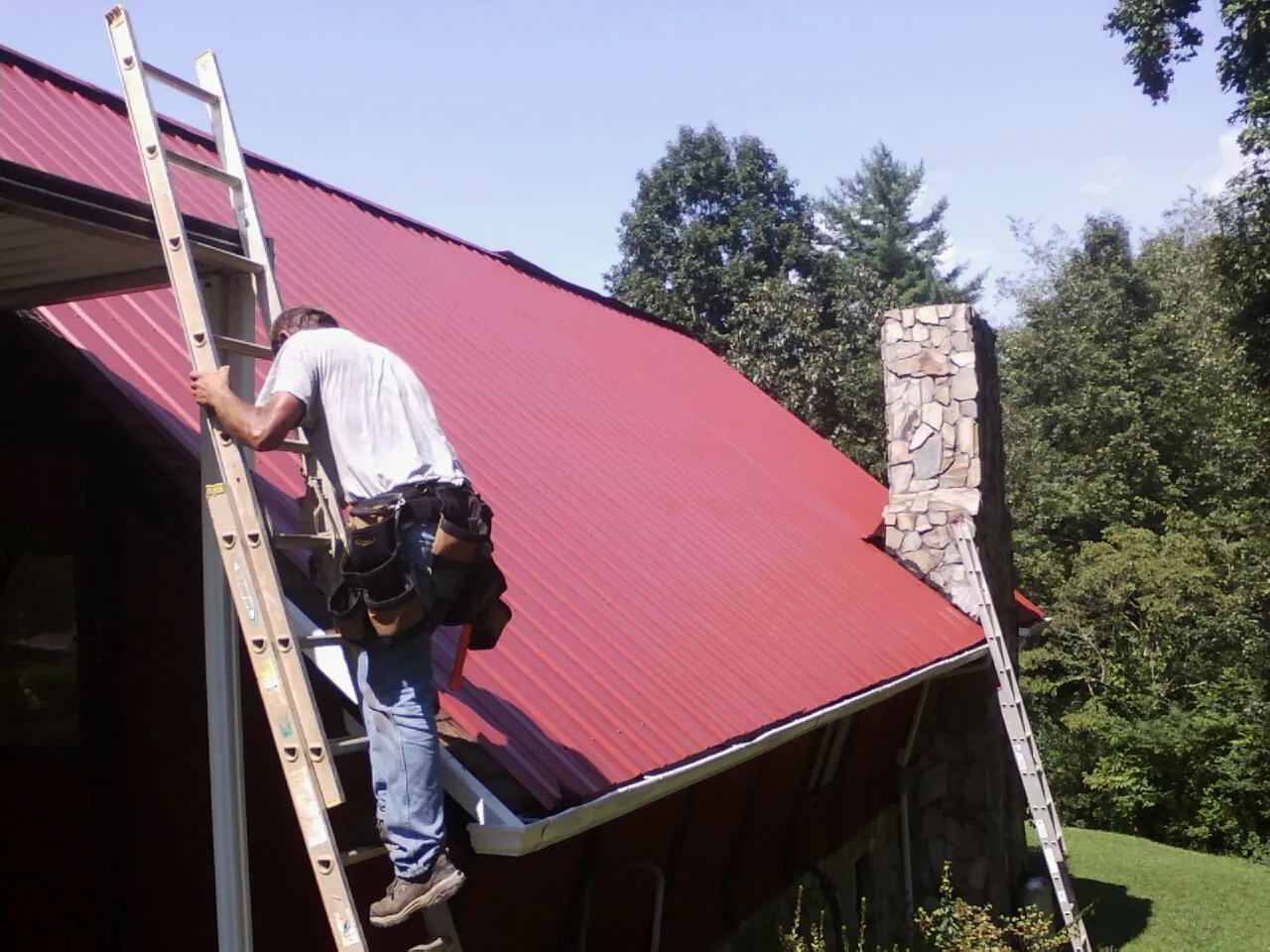 Roofer on a ladder beside a red metal roof and stone chimney — Soffit & Fascia Repair by Roofing Brothers in Newton Falls